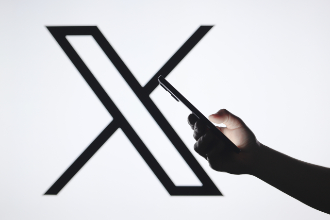 Một người dùng smartphone, phía sau là logo mạng xã hội X. Ảnh: Reuters
